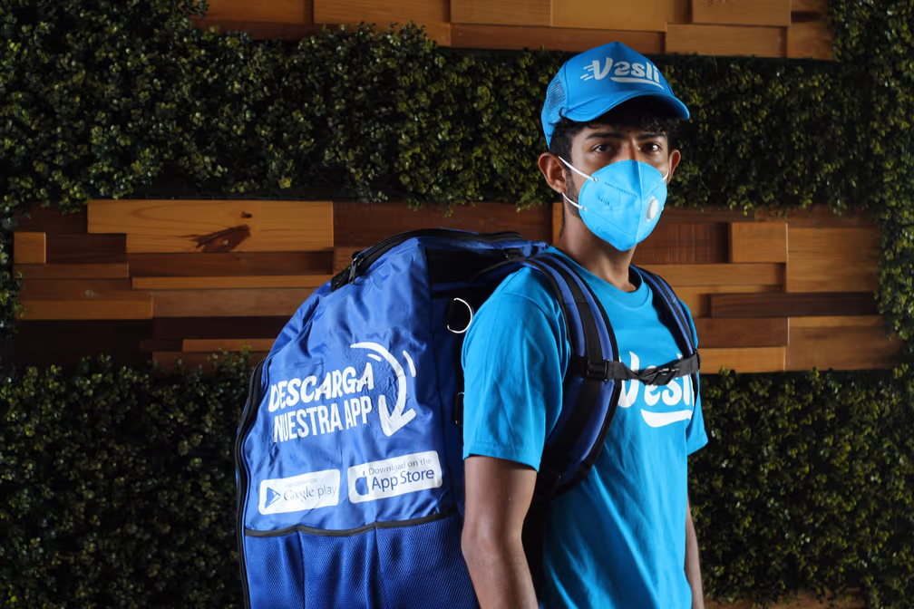 Socio repartidor de Vesli con la mochila puesta frente a un muro de madera y follaje